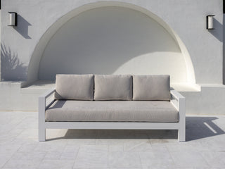 A modern outdoor 3 seater with a white frame and light beige cushions, positioned in front of a minimalist white stucco wall with a large arch.