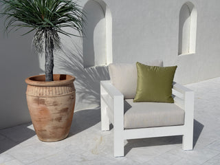 A modern white outdoor armchair with light beige cushions and a green accent pillow, positioned next to a large terracotta potted palm on a light stone patio against a white stucco wall with arched recesses.