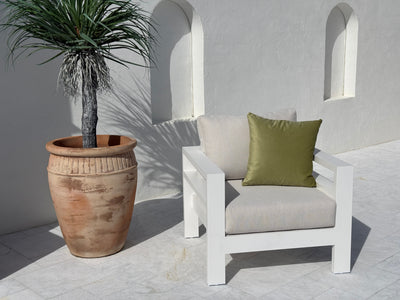 A modern white outdoor armchair with light beige cushions and a green accent pillow, positioned next to a large terracotta potted palm on a light stone patio against a white stucco wall with arched recesses.