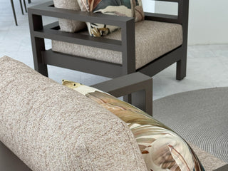 A close-up of a modern outdoor sofa with textured beige cushions and a decorative pillow, with a matching armchair in the background.