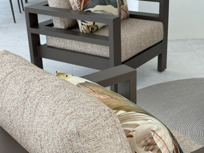 A close-up of a modern outdoor sofa with textured beige cushions and a decorative pillow, with a matching armchair in the background.