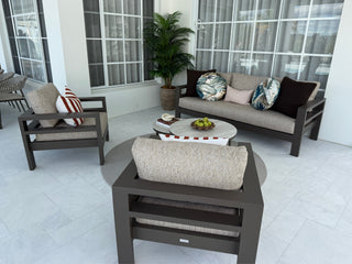 A modern outdoor patio conversation set featuring a dark brown frame with light beige textured cushions, including a three-seat sofa, two armchairs, and a round white coffee table, arranged on a light gray tiled patio.