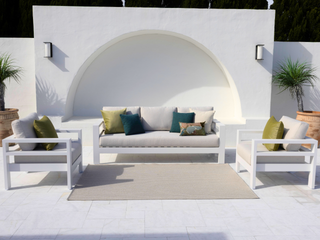 A modern outdoor patio furniture set featuring a white three-seat sofa, two matching armchairs, a beige area rug, and decorative throw pillows in olive green and teal, arranged in front of a white stucco wall with an arched recess.